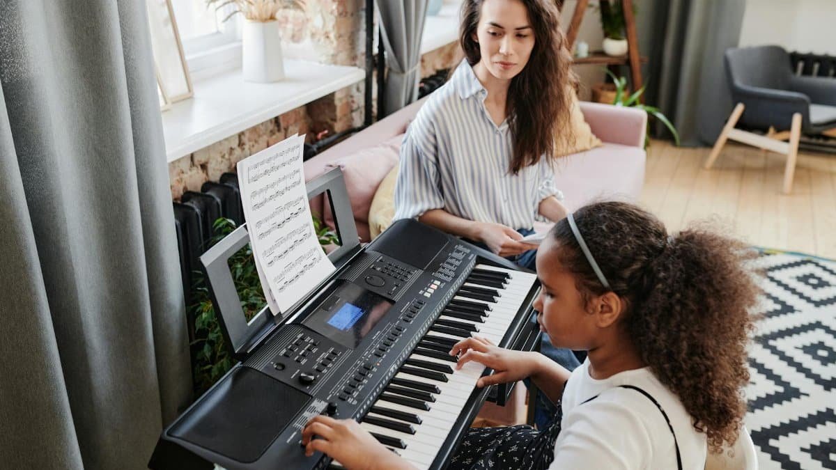 Girl takes piano lessons with attentive teacher observing indoors.