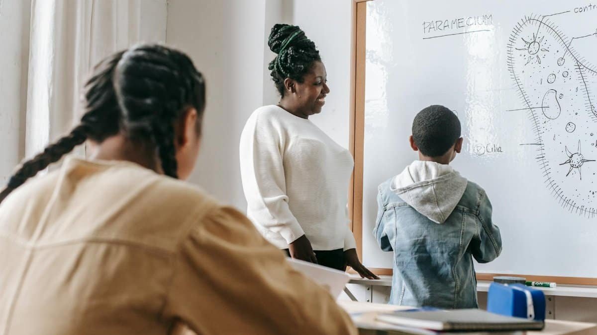 Smiling African American female teacher near white board with illustration and anonymous diverse schoolchildren in classroom