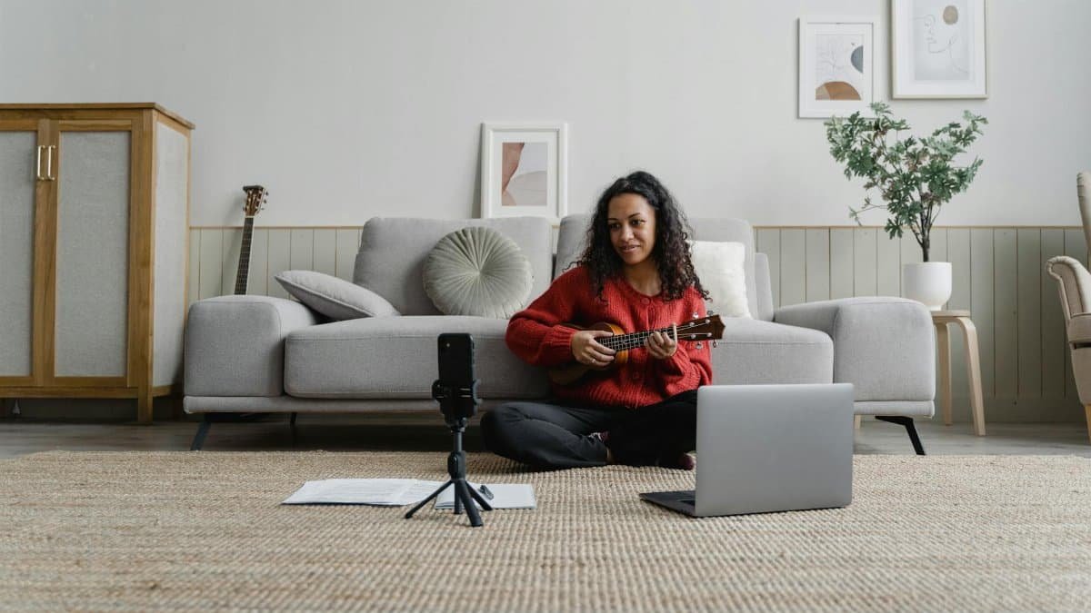 A woman sitting in her living room playing a ukulele during an online music lesson with a laptop and microphone.