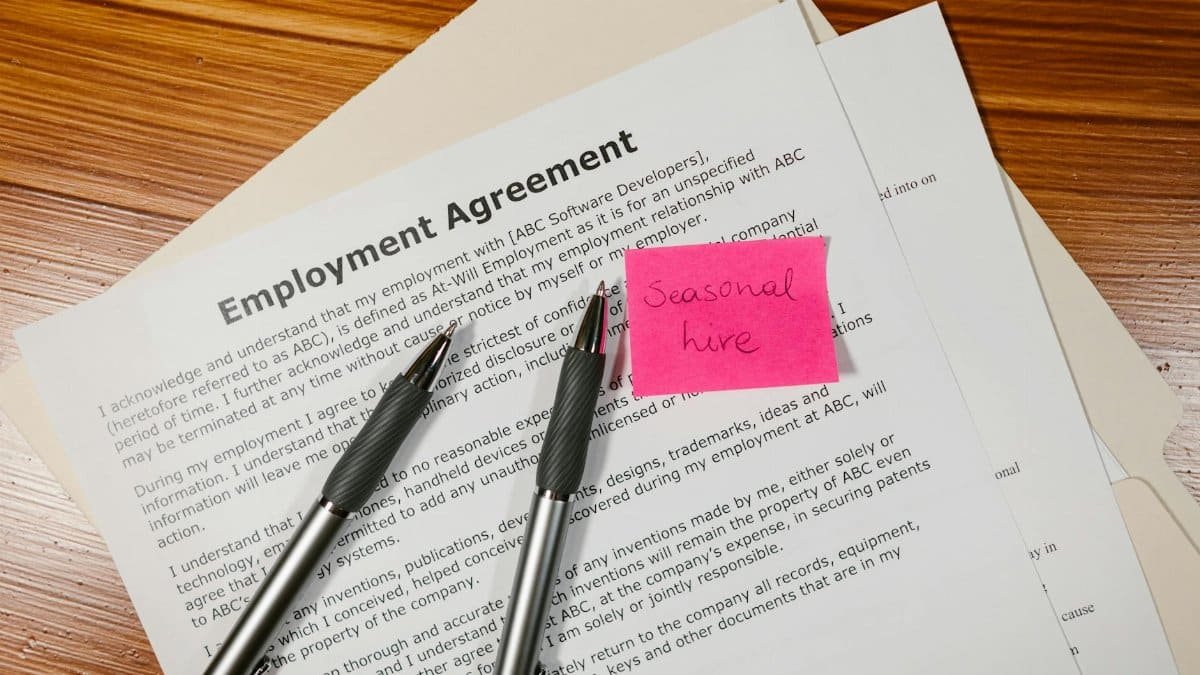 Top view of employment agreement contract with pens and seasonal hire note on wooden desk.