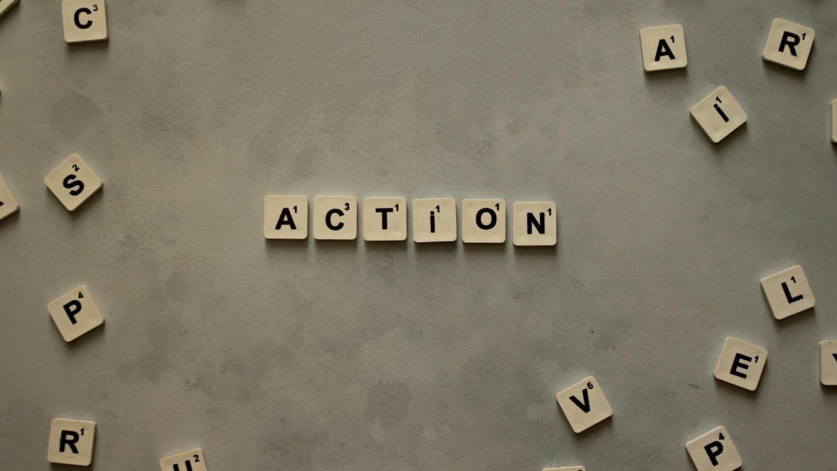 Flat lay of Scrabble tiles spelling 'Action' on a gray background, surrounded by random letters.