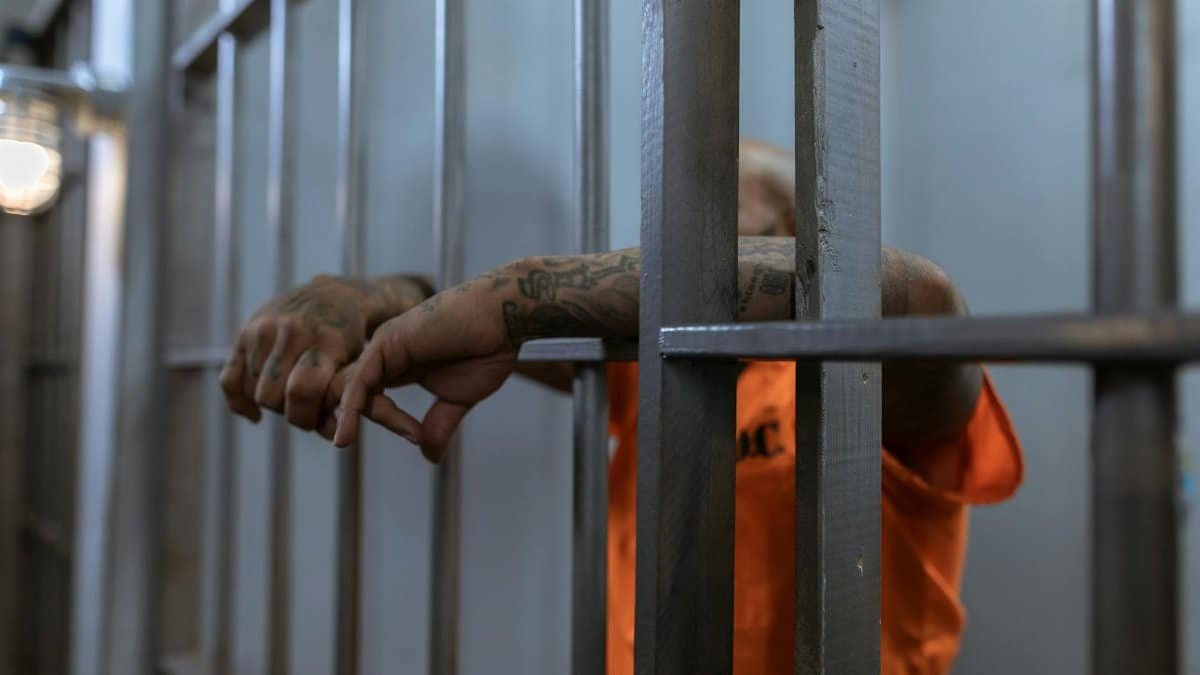A tattooed inmate's arms rest on jail cell bars, wearing an orange uniform inside a prison.