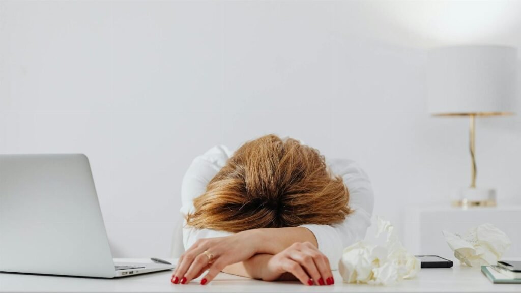 Tired woman resting head on arms at desk with laptop, showing fatigue in modern office.