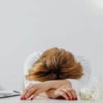Tired woman resting head on arms at desk with laptop, showing fatigue in modern office.