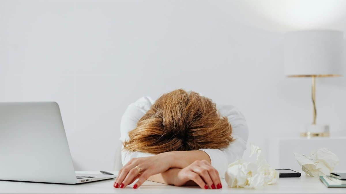 Tired woman resting head on arms at desk with laptop, showing fatigue in modern office.