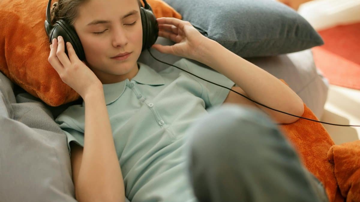 A young girl with closed eyes enjoys music through wired headphones while reclining indoors.