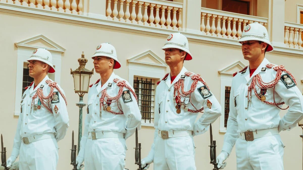 Four uniformed guards in white attire stand in formation outside a classical building.