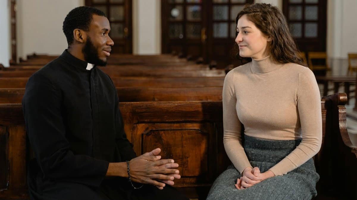 A priest and a woman seated on wooden pews in discussion, symbolizing faith and community.