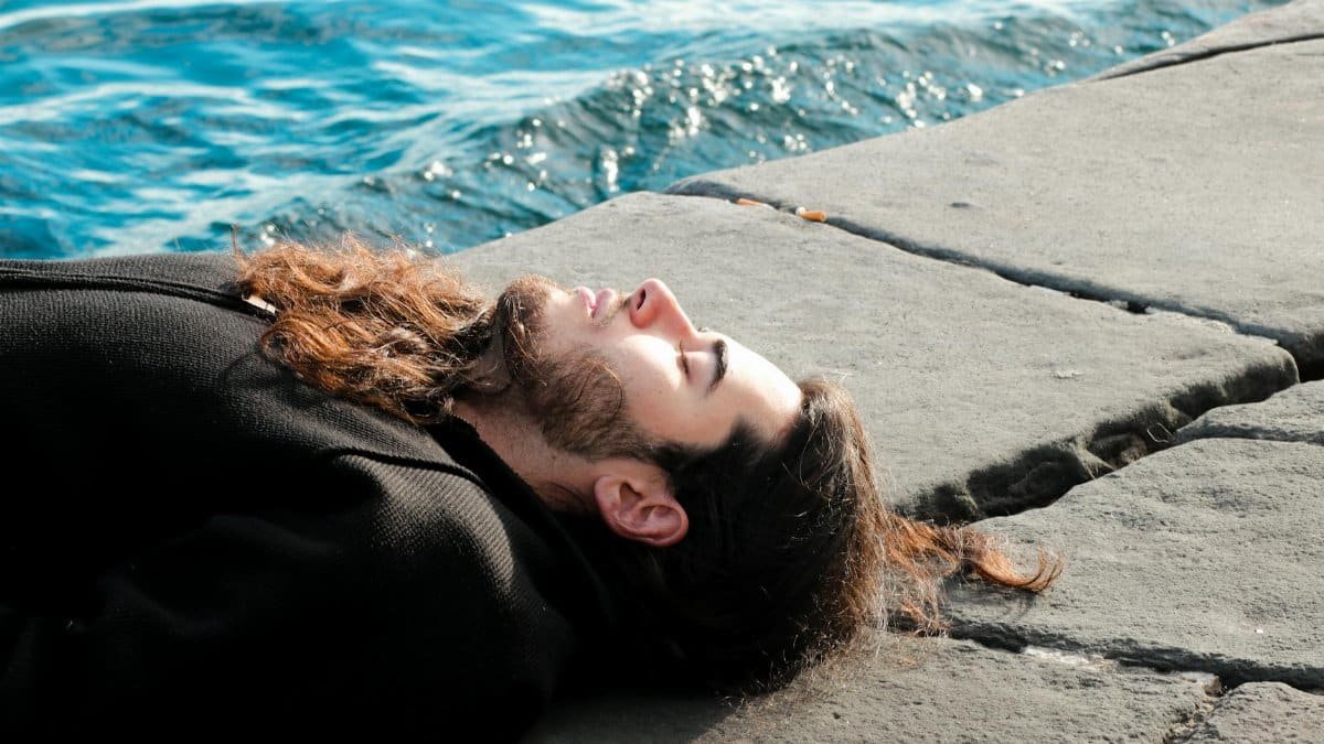 A man with long hair and beard lying by the ocean, enjoying a sunny and relaxing day.