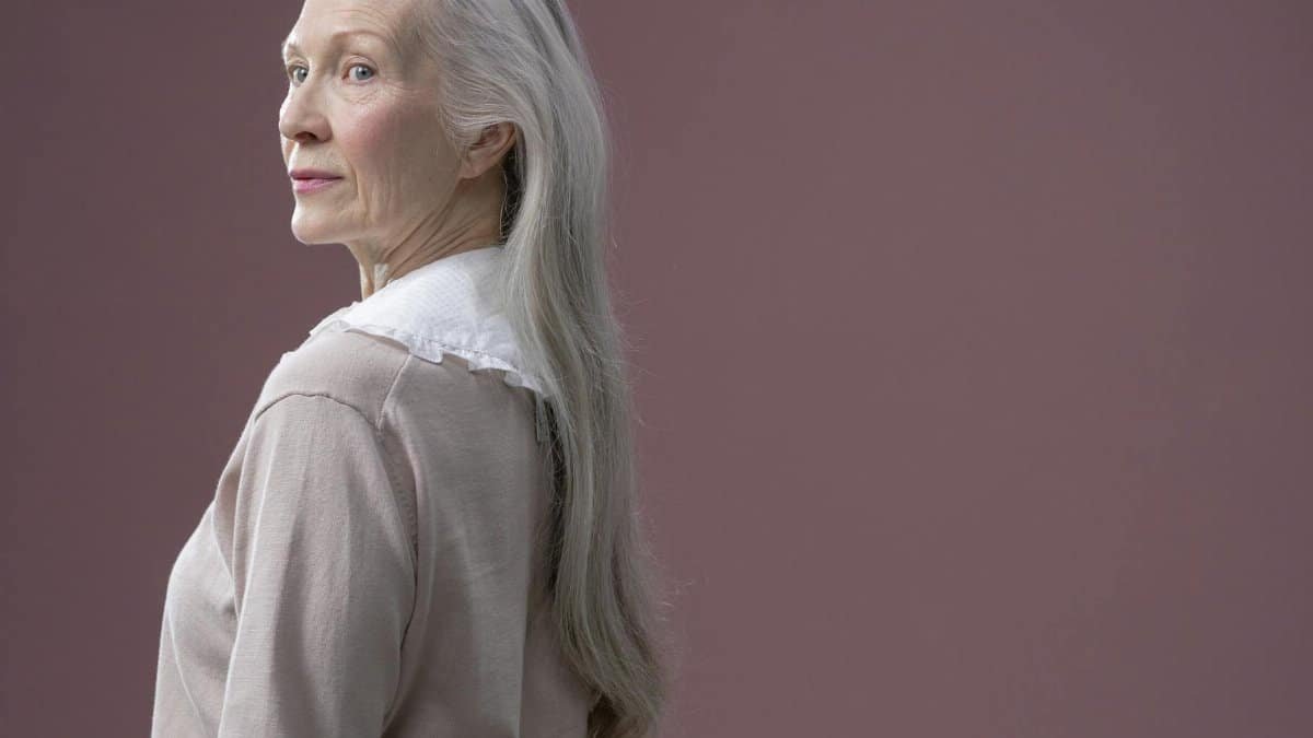 A sophisticated senior woman with long gray hair stands in profile against a solid background.