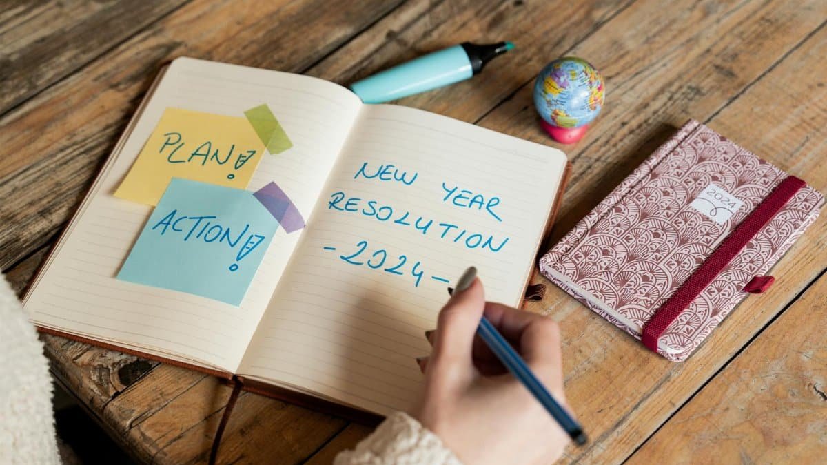 Person writing New Year 2024 resolutions in a notebook with sticky notes for planning and action.