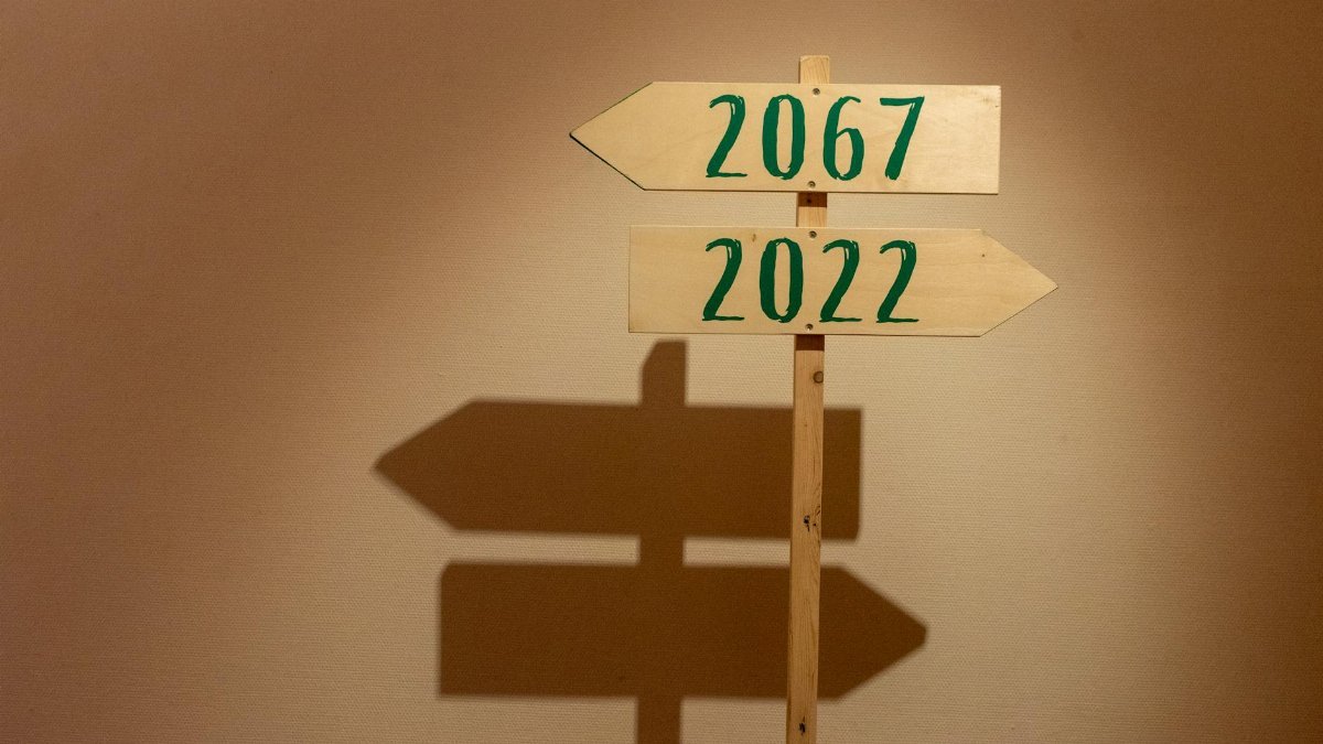 Wooden signpost indoors pointing to years 2067 and 2022, casting shadows.