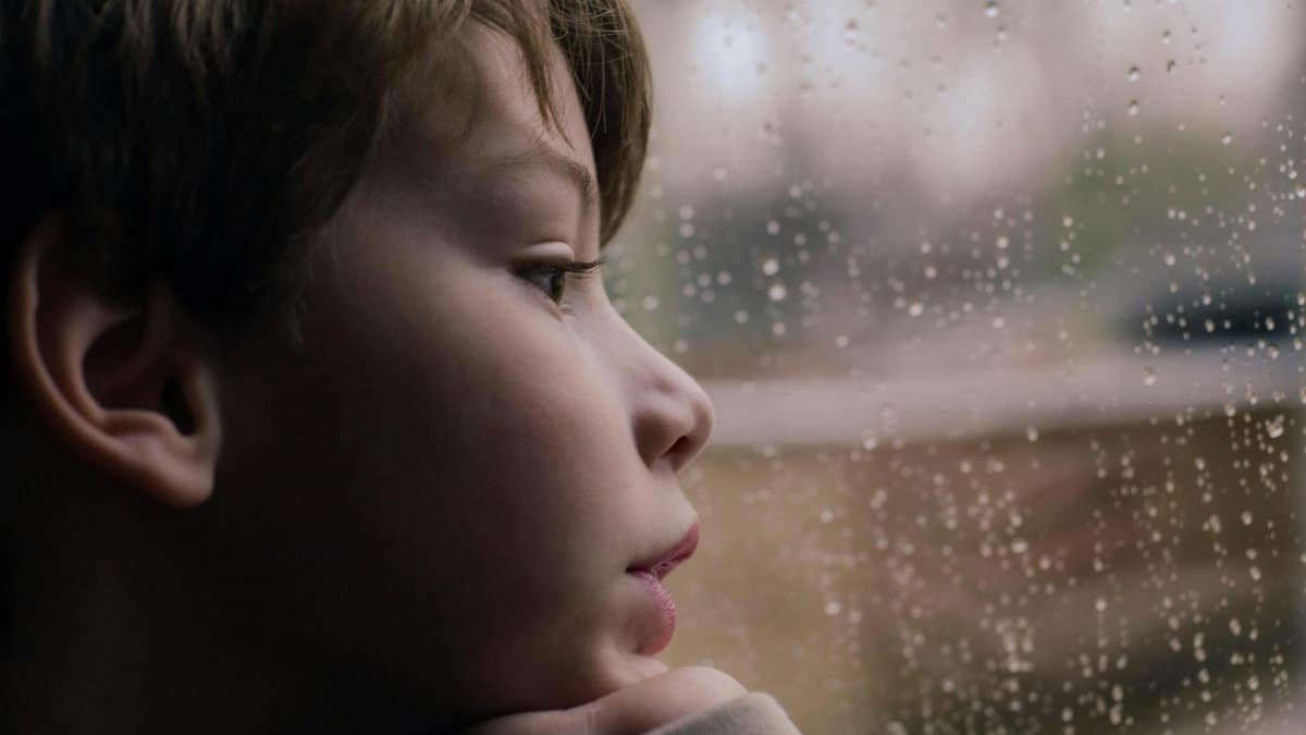 Contemplative child gazes through rain-speckled window, evoking a sense of calm and reflection.