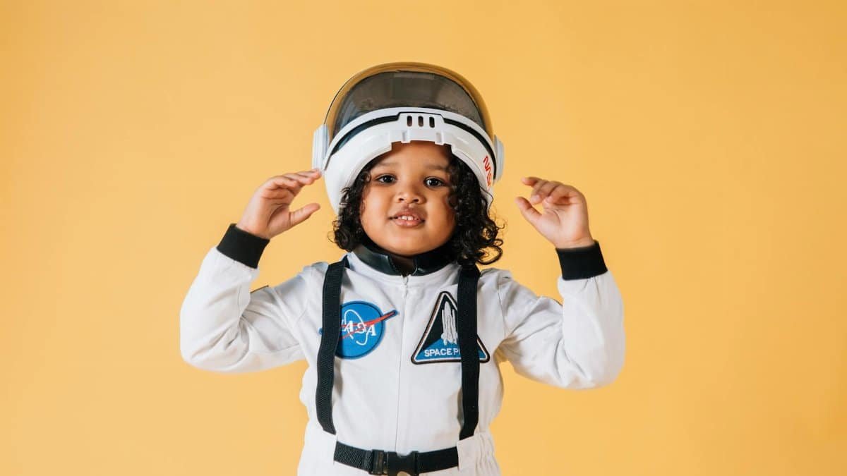 Cheerful little African American girl wearing white astronaut spacesuit and helmet and looking at camera contentedly while standing against brown wall
