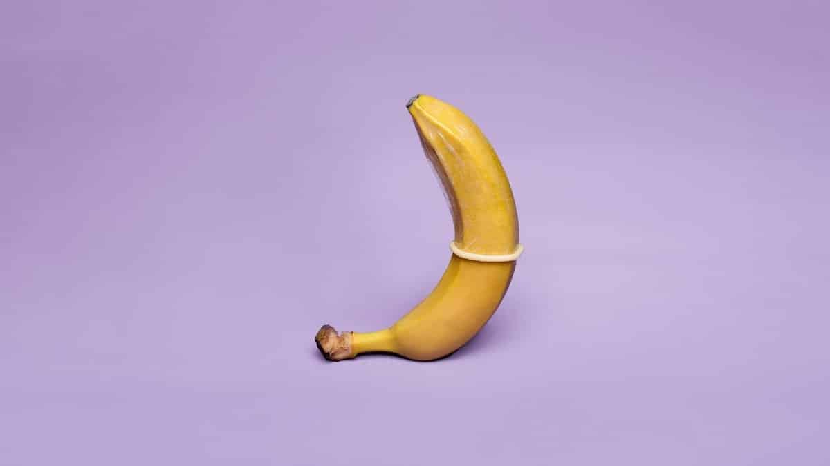 A banana wearing a condom symbolizes safe sex education against a pastel purple background.