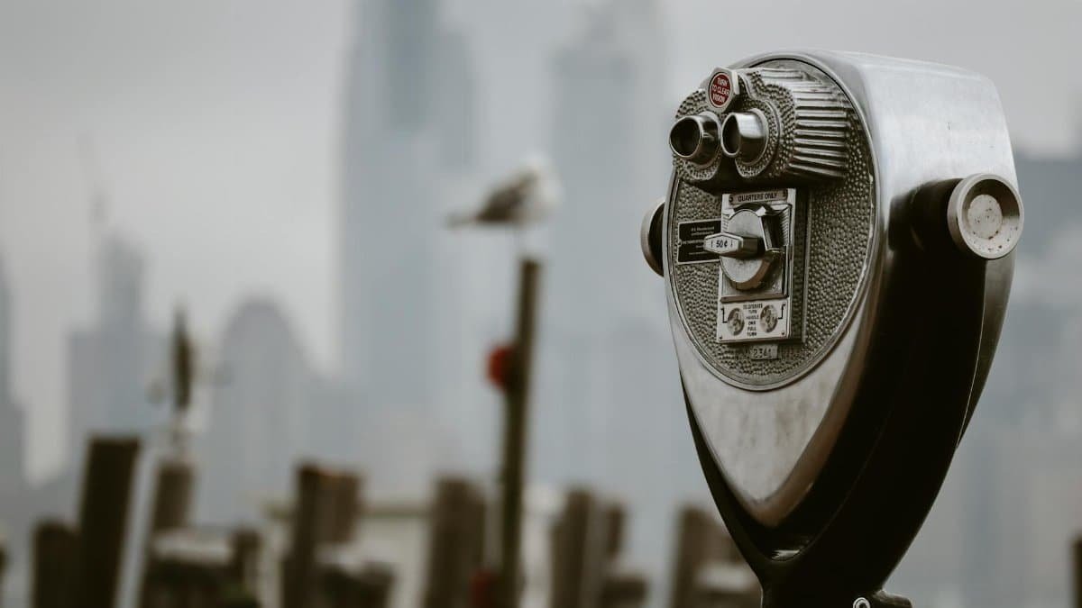 A coin-operated binocular viewer overlooks a blurred New York City skyline on a cloudy day.