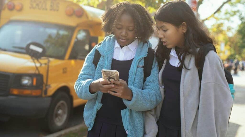 Content multiracial girls classmates wearing warm jackets browsing modern mobile phone while standing together on sunny autumn street near school bus