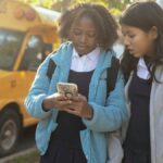 Content multiracial girls classmates wearing warm jackets browsing modern mobile phone while standing together on sunny autumn street near school bus