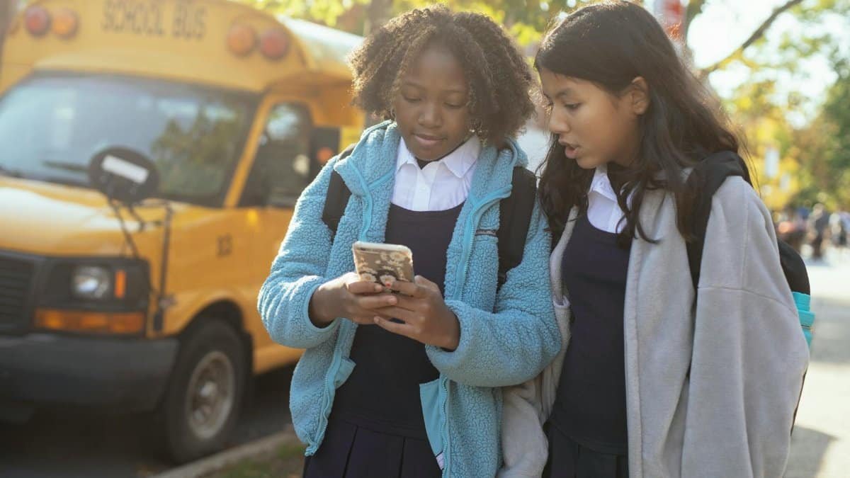 Content multiracial girls classmates wearing warm jackets browsing modern mobile phone while standing together on sunny autumn street near school bus