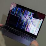 A broken laptop screen displayed with colorful glitch being held by a person.