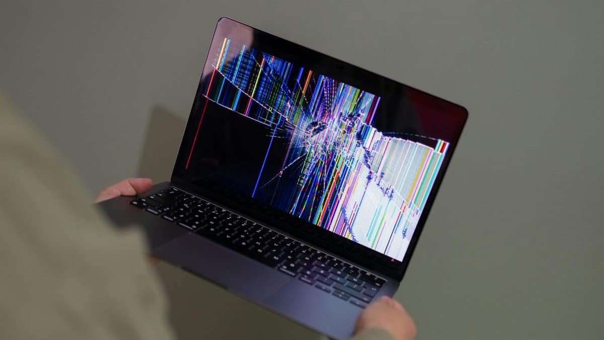 A broken laptop screen displayed with colorful glitch being held by a person.