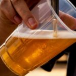 Close-up of a person pouring golden craft beer into a clear plastic cup.