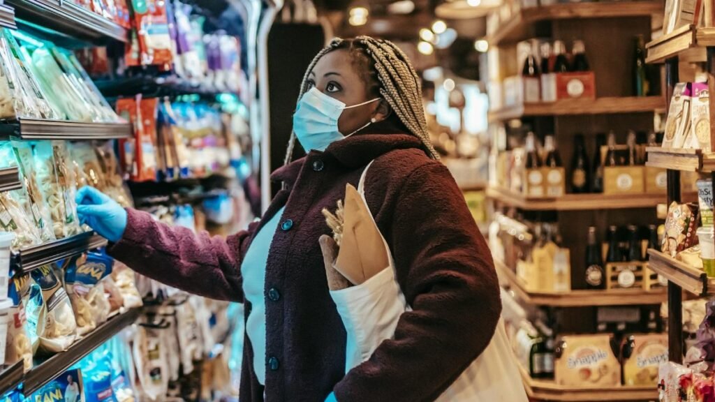 Side view of African American female shopper in protective mask and gloves selecting goods in grocery store