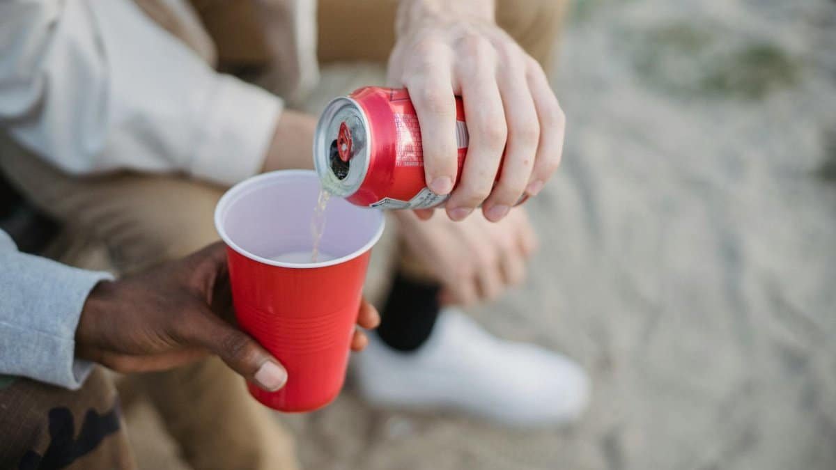 From above of crop anonymous man pouring fizzy drink from can into red plastic cup of black friend while sitting on sandy ground