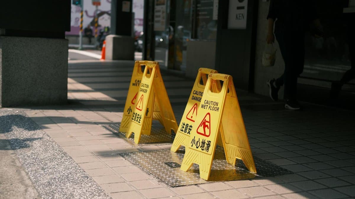 Yellow caution wet floor signs on a sunlit outdoor sidewalk, indicating a safety warning.