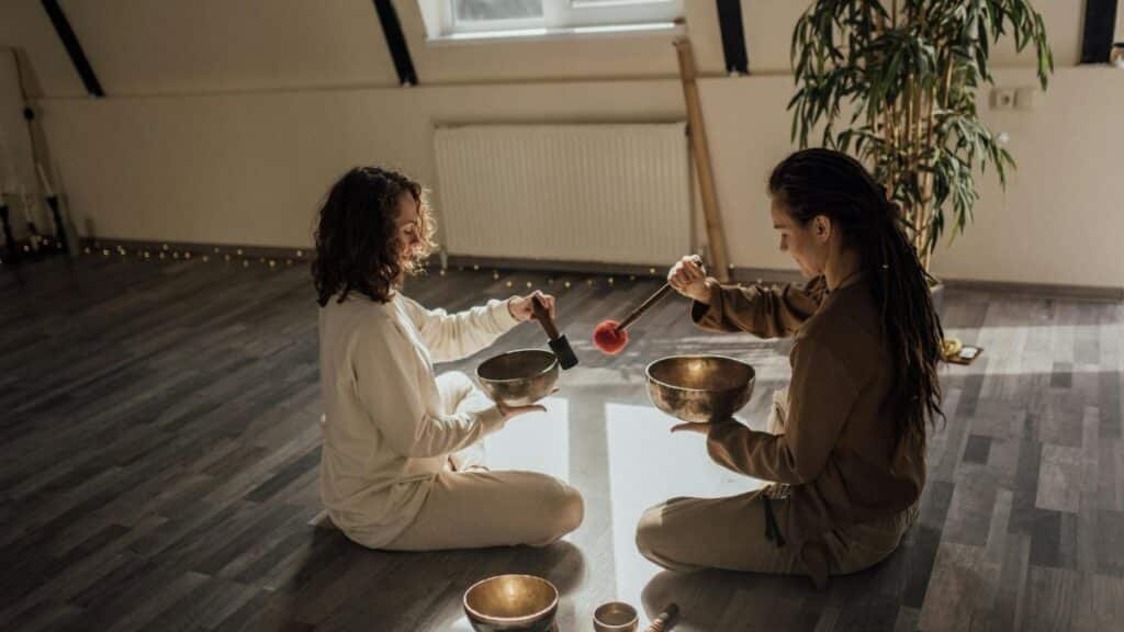 Two women sitting and meditating with Tibetan singing bowls indoors, creating a serene atmosphere.