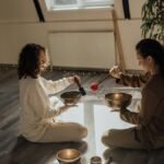 Two women sitting and meditating with Tibetan singing bowls indoors, creating a serene atmosphere.