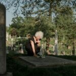 An elderly woman in a black dress mourns at a grave in a peaceful cemetery setting.