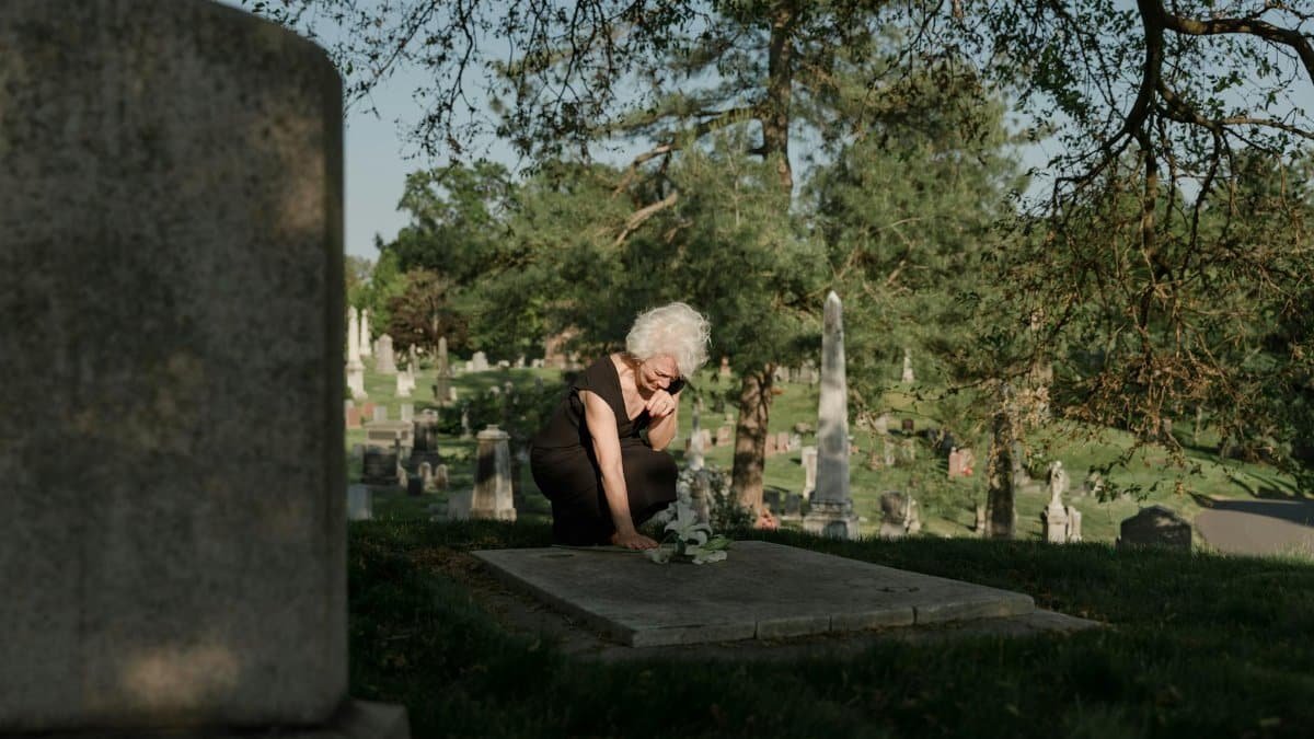 An elderly woman in a black dress mourns at a grave in a peaceful cemetery setting.