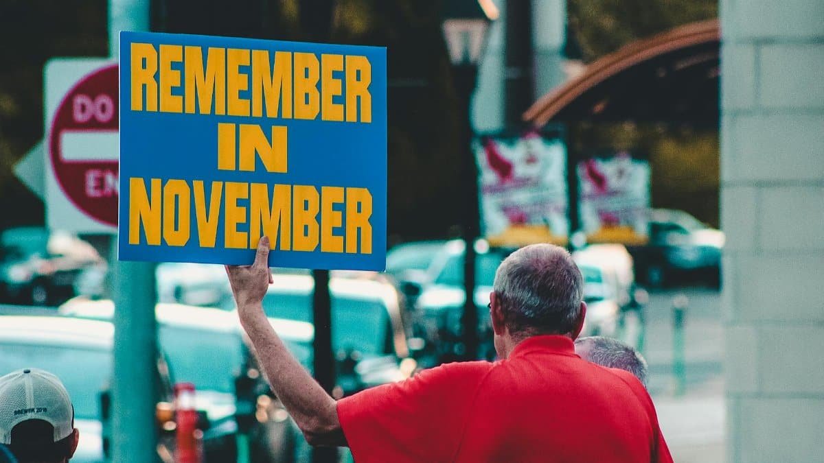 Man holding 'Remember in November' sign on urban street during the day.
