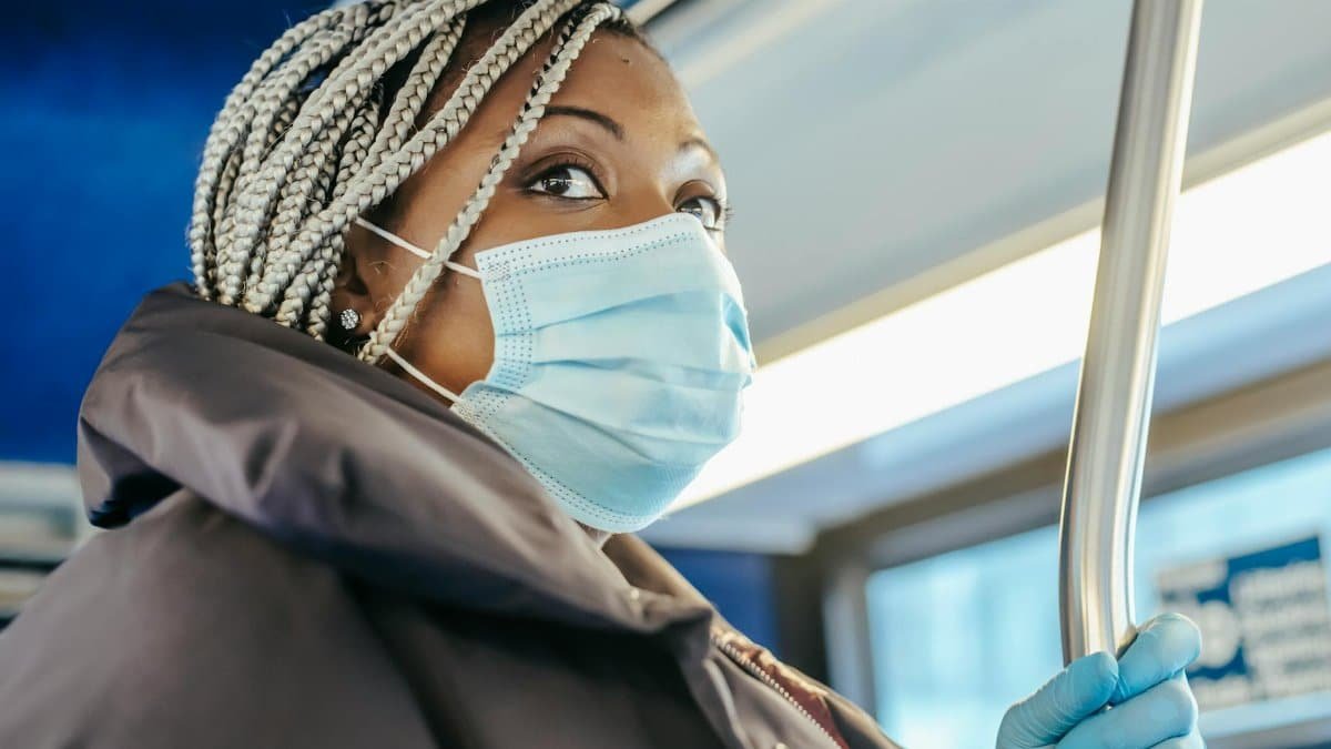 Adult black woman in warm outfit and protective mask with gloves riding on public bus in daytime