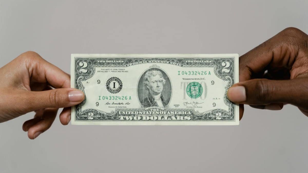 Close-up of two hands exchanging a two-dollar bill on a neutral background.