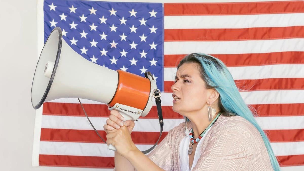 Expressive woman using a megaphone in front of a vibrant American flag, symbolizing freedom.