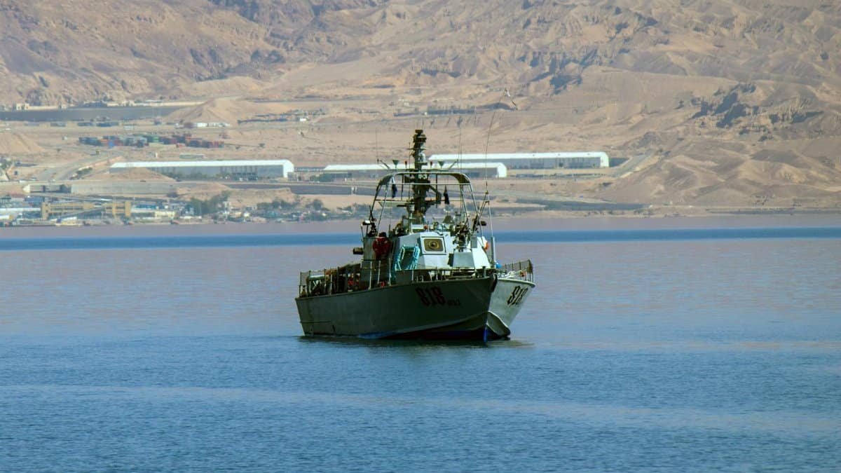A naval patrol boat sails through the deep blue water with a desert landscape in the background.