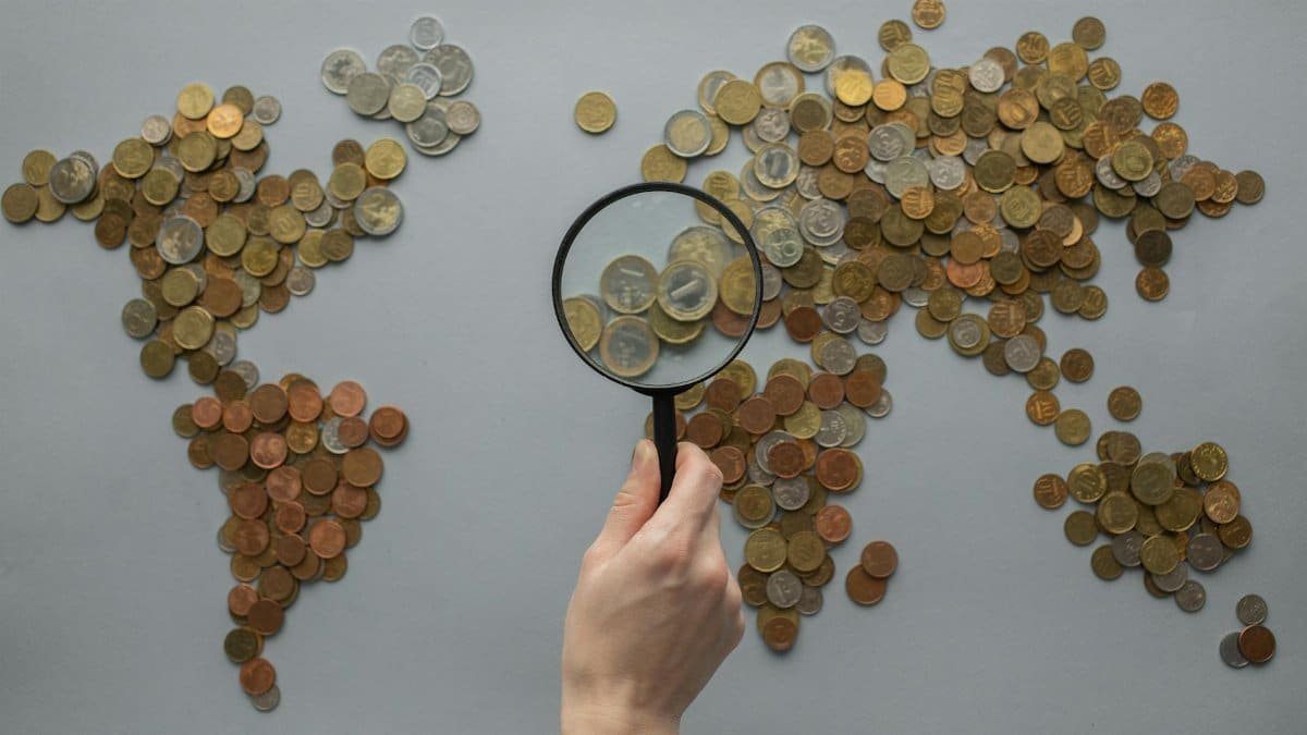 Top view of crop unrecognizable traveler looking through magnifying glass on world map made of various coins on gray background