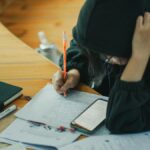 A student wearing a hoodie is deeply engaged in writing notes at a wooden study desk, surrounded by educational materials.