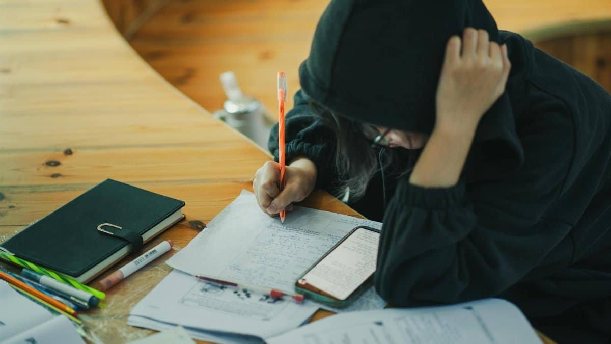 A student wearing a hoodie is deeply engaged in writing notes at a wooden study desk, surrounded by educational materials.