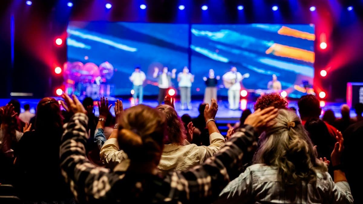 Audience members raise hands at a live concert with colorful stage lighting and performers.