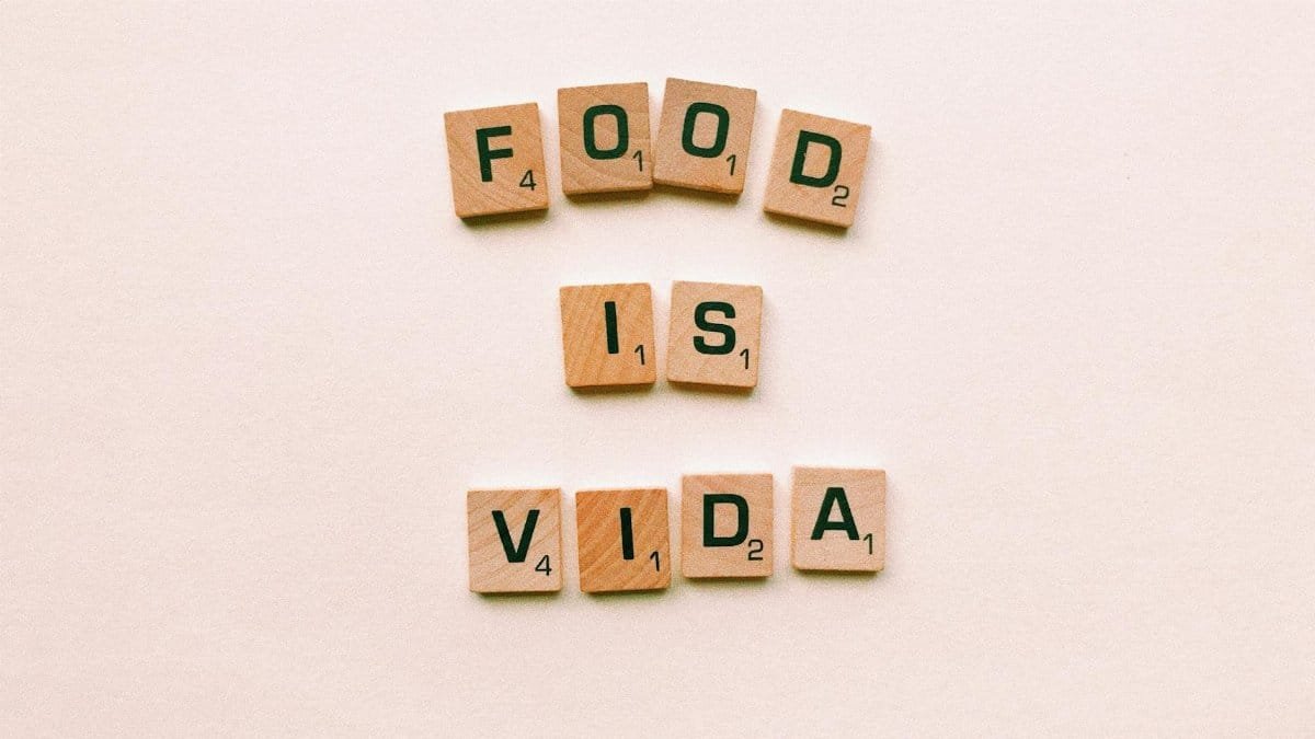 Wooden letter tiles spell 'FOOD IS VIDA' on a soft pink background.