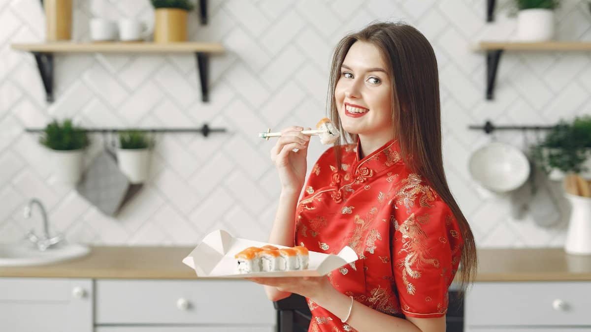 Happy young female with long dark hair in vivid dress enjoying Japanese traditional cuisine and eating appetizing sushi while having lunch in modern light kitchen