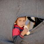 A young boy peering through a torn tent in Idlib, reflecting innocence amid hardship.