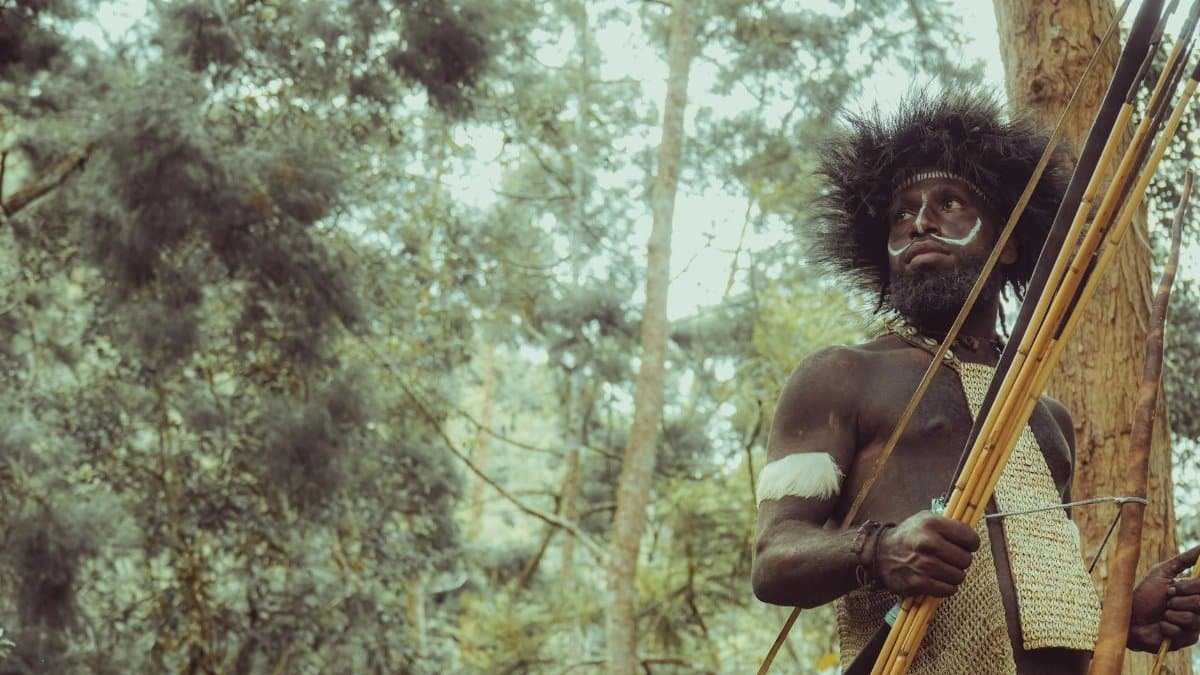 A tribal man with traditional attire and face paint holds spears in a forest environment.