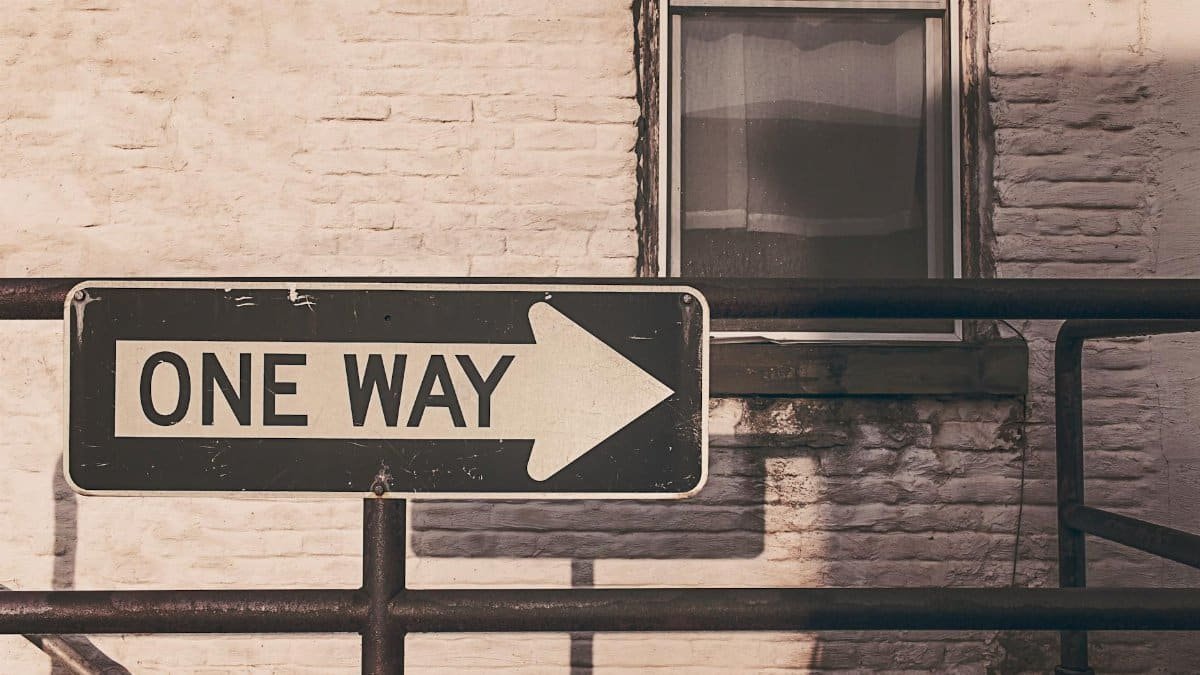 A vintage one way sign pointing right, mounted on a metal railing against a brick wall in urban setting.