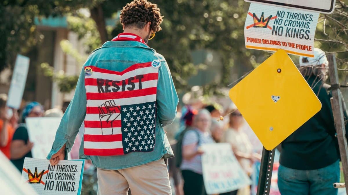 Person wearing flag-themed denim jacket at a protest holding signs promoting resistance and anti-establishment views.