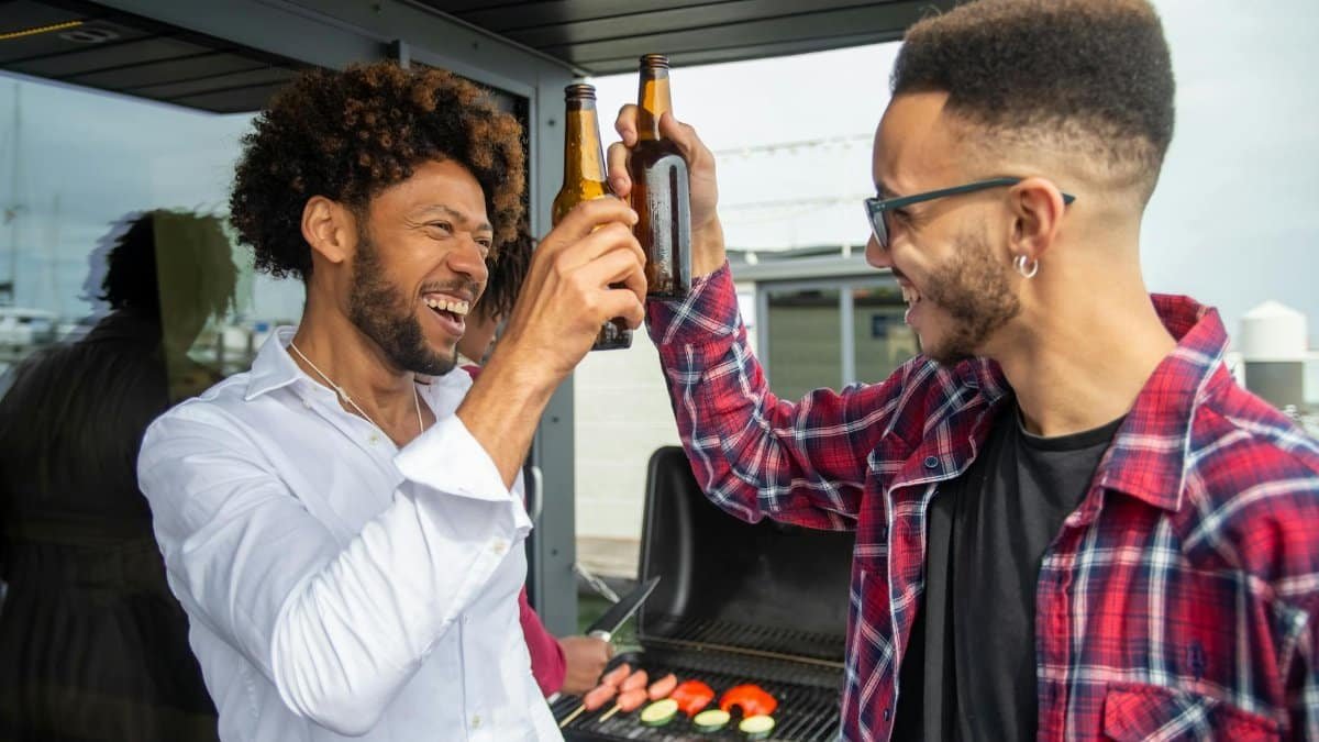 Two friends clink beer bottles while cooking at an outdoor barbecue in Portugal.