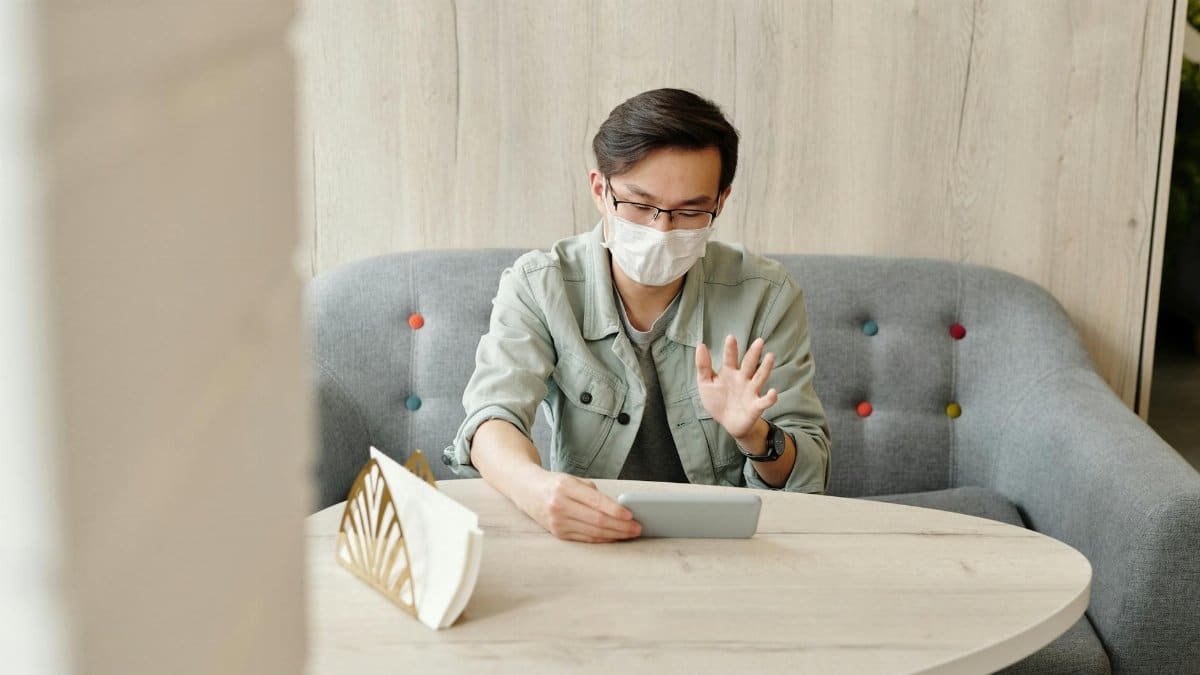 Man wearing a mask while using a tablet for a video call indoors during daytime.